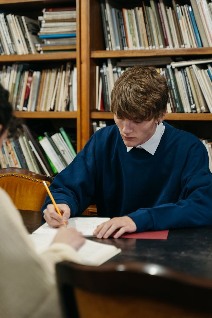 A young man in a blue sweater writing on paper in a library setting.